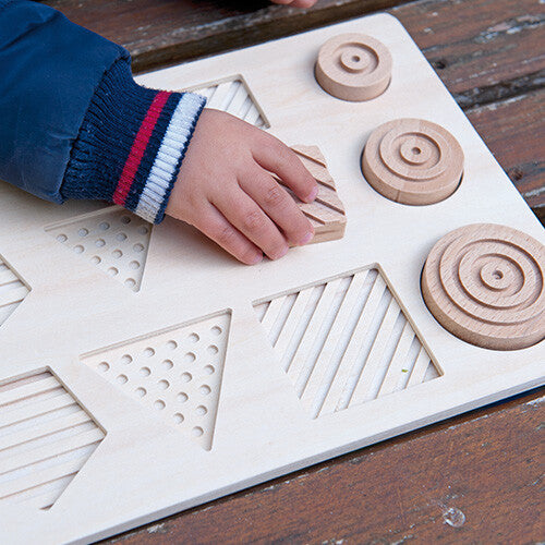 Een kinderhand plaatst een vierkant puzzelstuk met streepjestextuur in de Andreu Toys houten sensorische puzzel.
