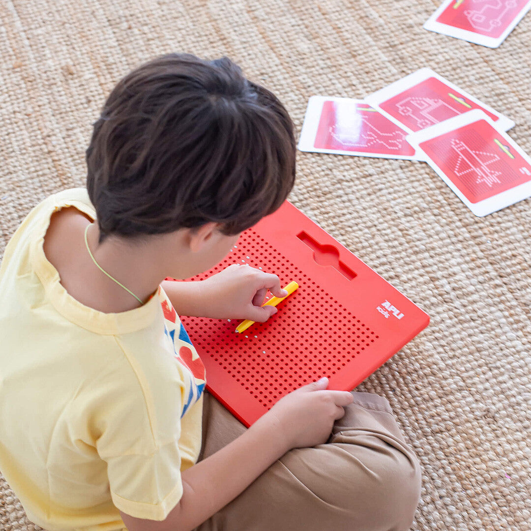 Kind tekent op een rood APLI kids magnetisch tekenbord XL met gele stylus, met patroonkaarten op de achtergrond.