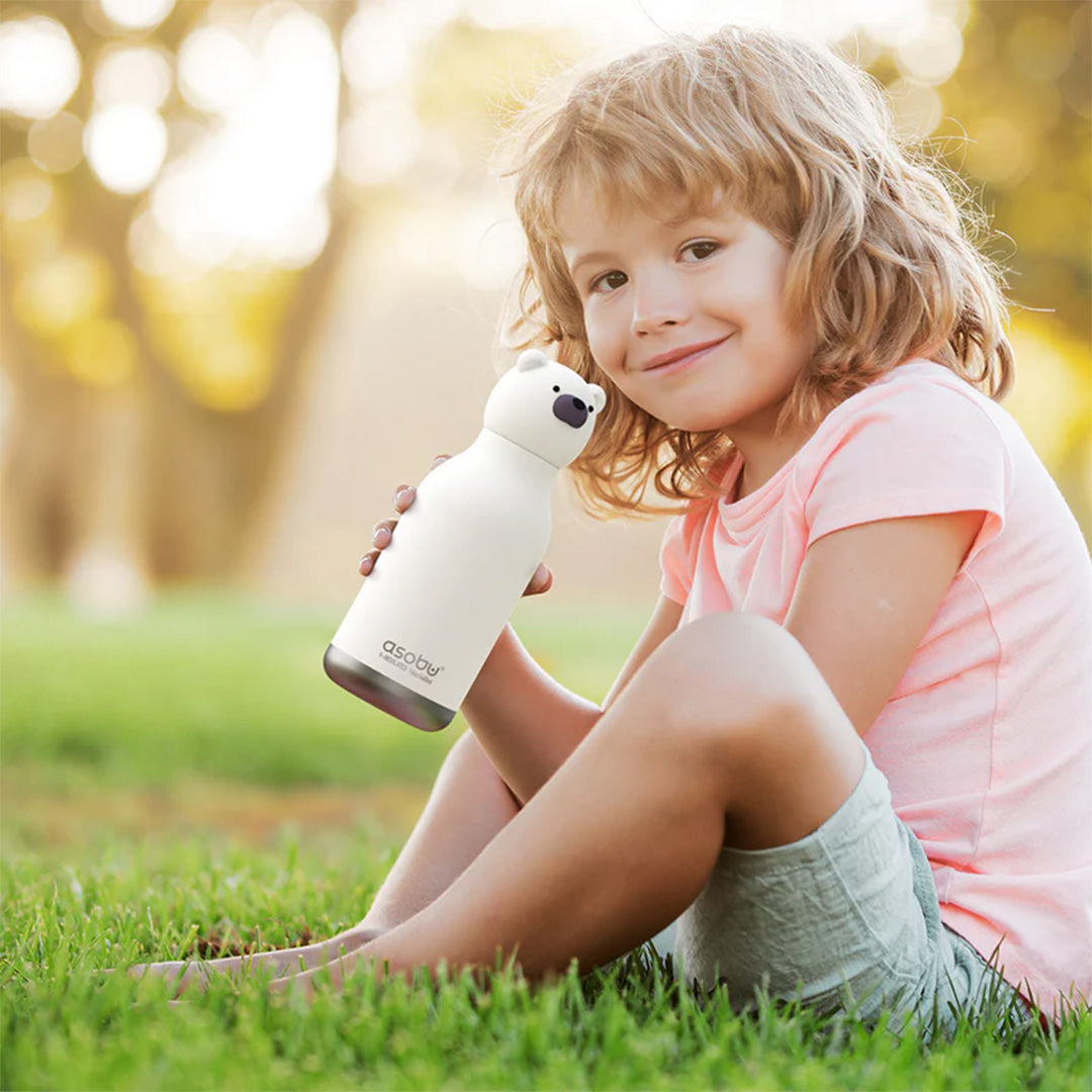 Een lachend kind zit op het gras en houdt een asobu rvs drinkfles in de vorm van een beer vast.
