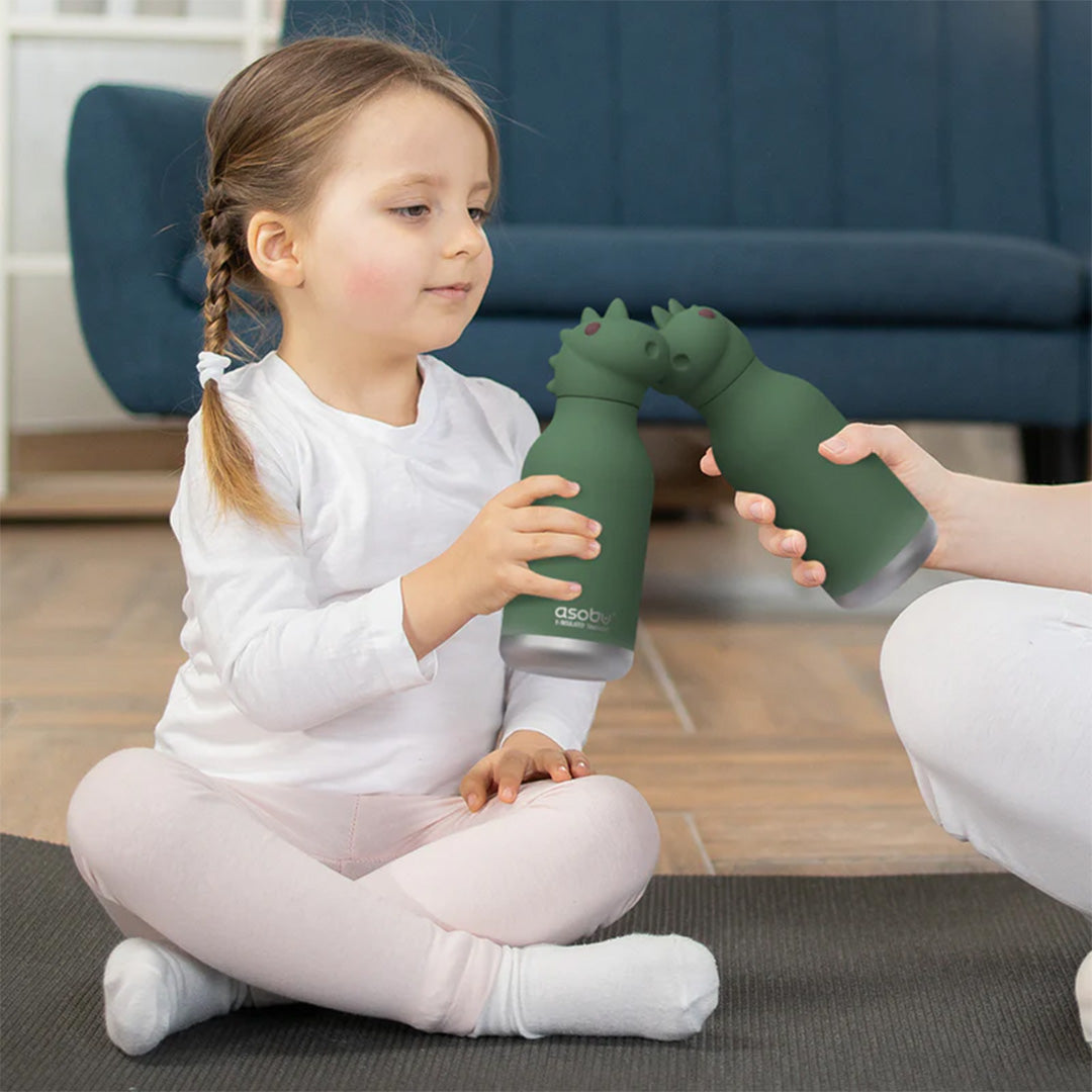 Een jong meisje houdt een groene asobu dinosaurus drinkfles vast, terwijl een volwassene haar een tweede fles aanreikt.