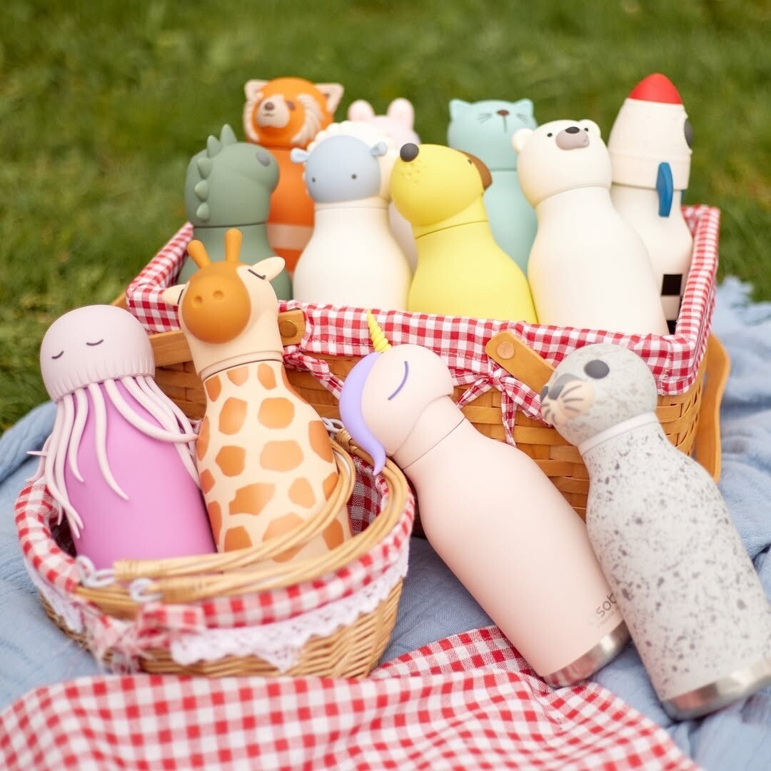 Collectie asobu Bestie drinkflessen met diverse dierenfiguren in een picknickmand op een rood-wit geruite deken.