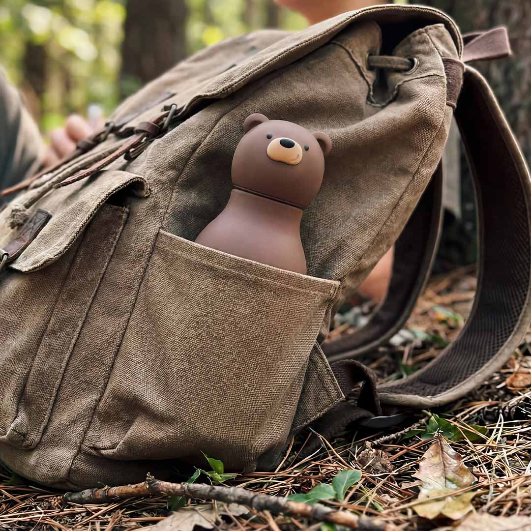 De asobu drinkfles in de vorm van een bruine beer steekt uit de zijzak van een rugzak in het bos.