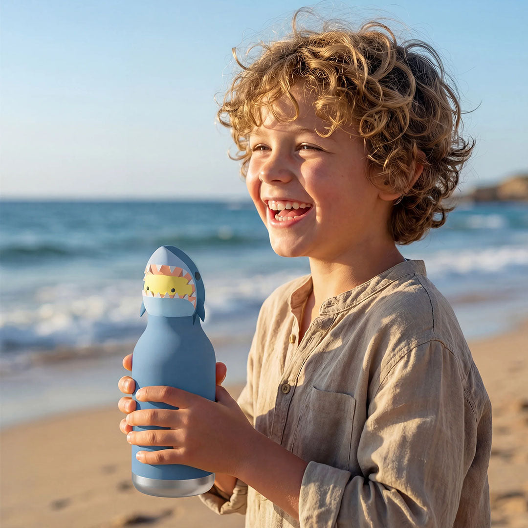 Een lachende jongen houdt de asobu haai drinkfles vast op een zandstrand met de zee op de achtergrond.