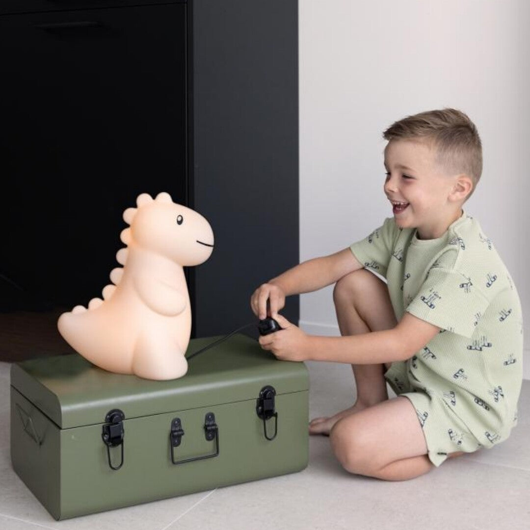 Een lachend kind knielt naast de zandkleurige dino kinderlamp die op een groene kist staat.