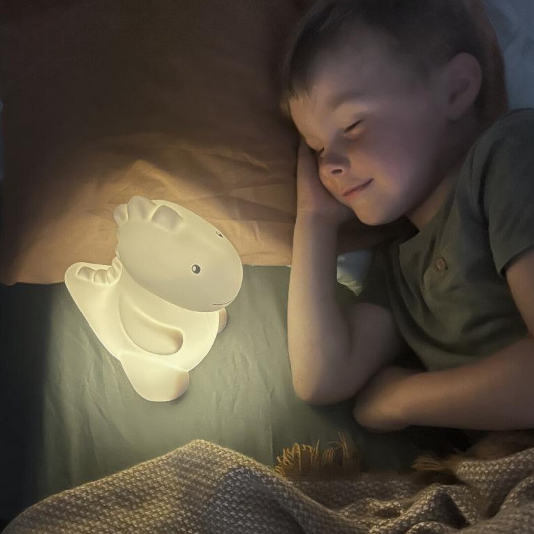 Een slapende jongen met het zacht gloeiende atelier pierre dinosaurus LED nachtlampje naast zijn hoofd in bed.