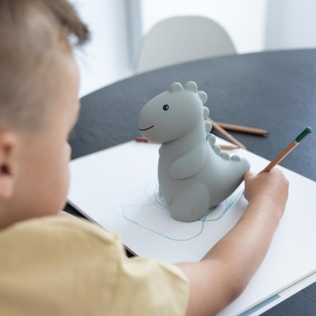 Een kind tekent aan tafel met de Atelier Pierre groene dinosaurus spaarpot ernaast.