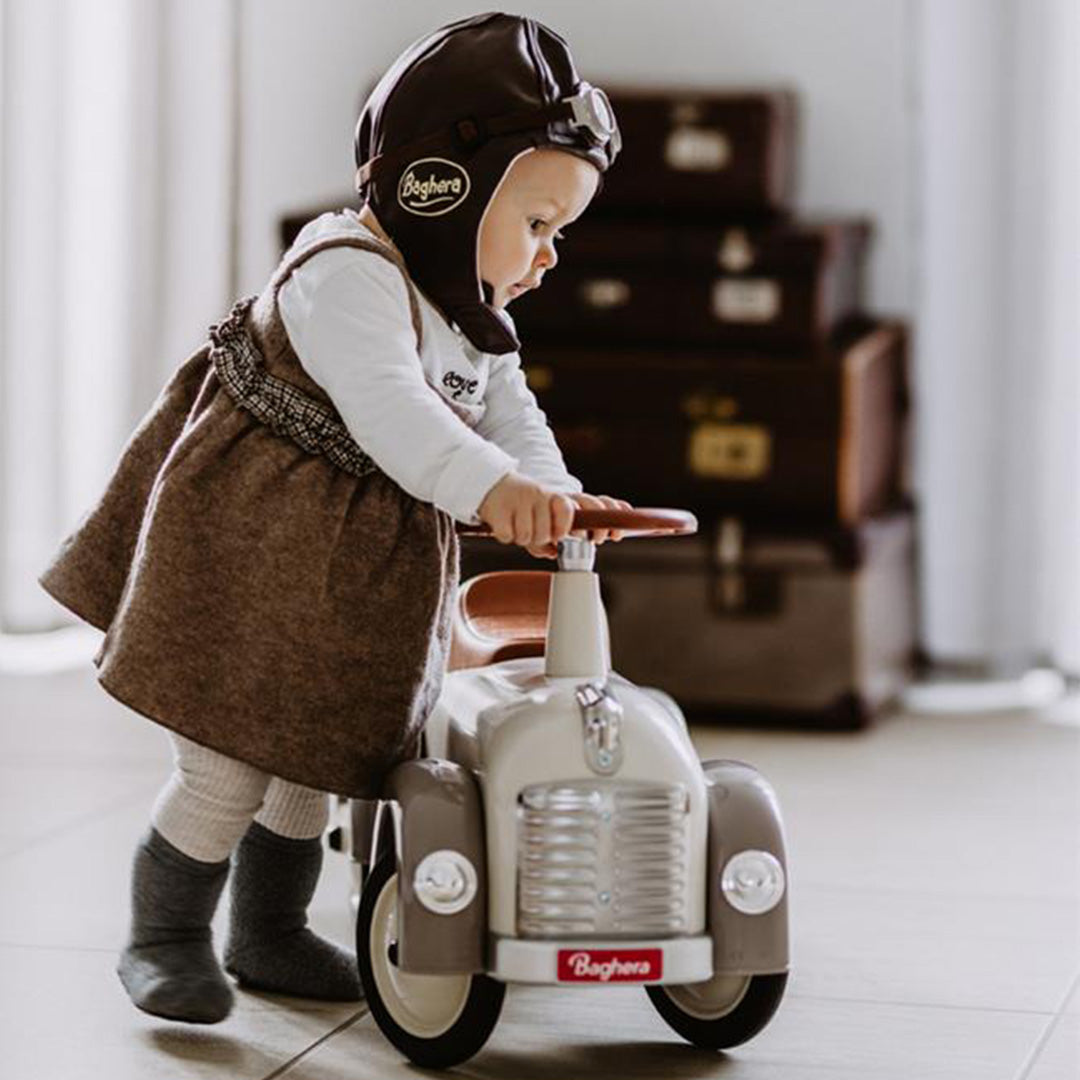 Peuter met pilotenmuts staat bij de voorkant van de Baghera loopauto, beige met grijze details.