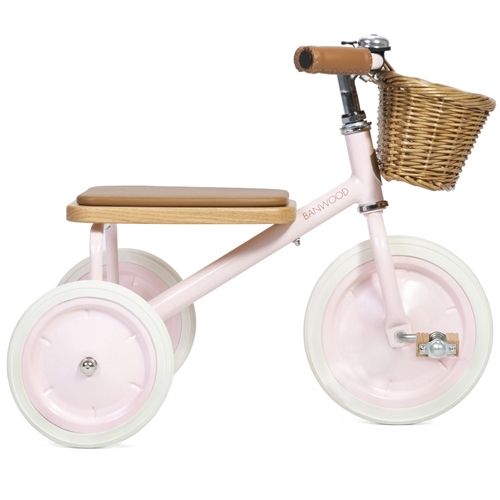 Banwood roze driewieler met houten zitting en rotan mandje, schuin van opzij gefotografeerd.