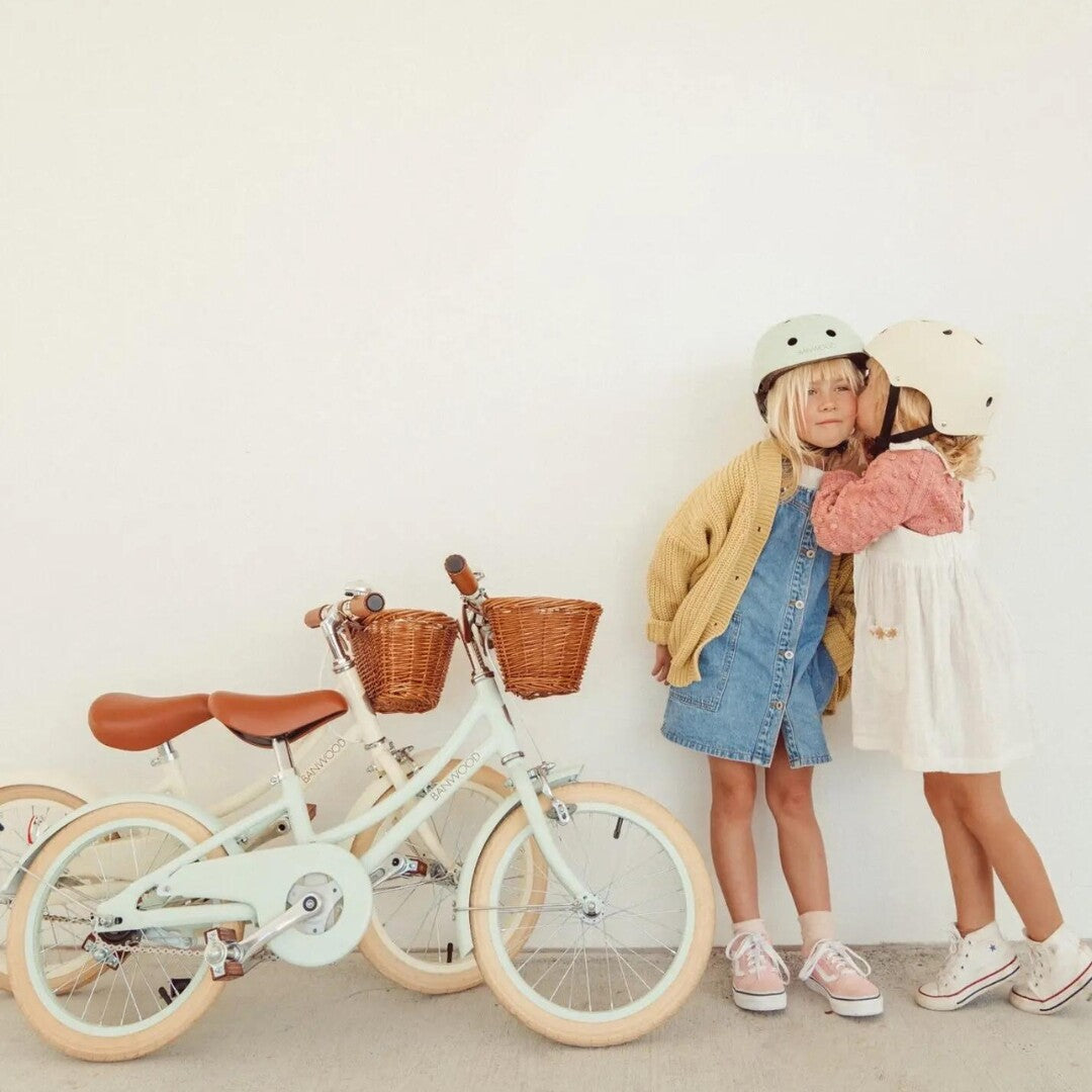 Twee kinderen met helmen knuffelen naast twee Banwood kinderfietsen, één crèmekleurig en één mintgroen.