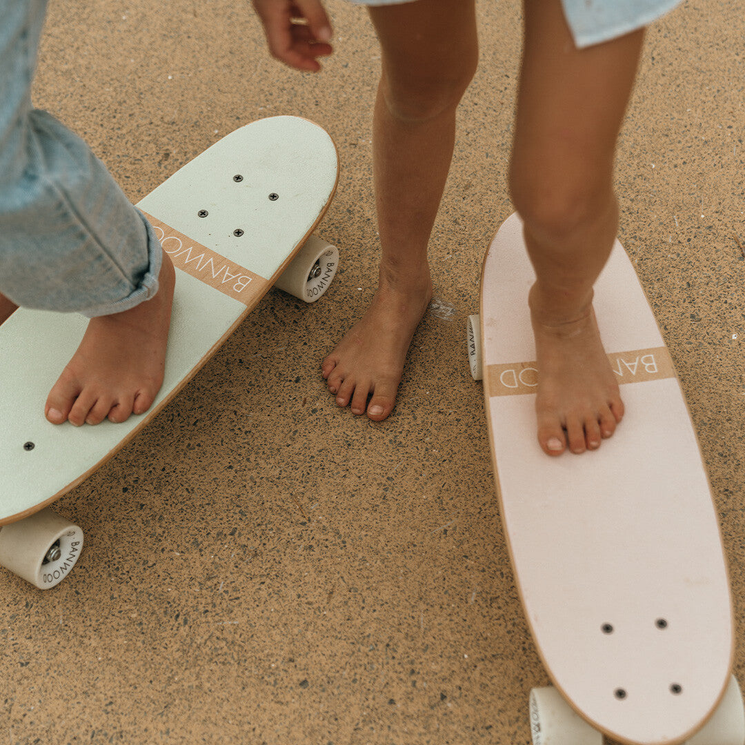 Twee kinderen met blote voeten op twee Banwood kinderskateboards op een asfaltweg.