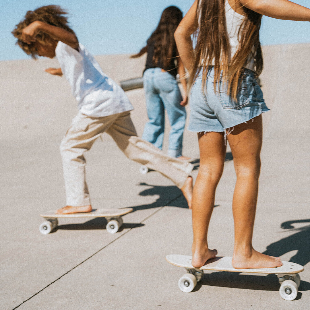 Drie kinderen rijden blootsvoets op Banwood kinderskateboards op een betonnen ondergrond.