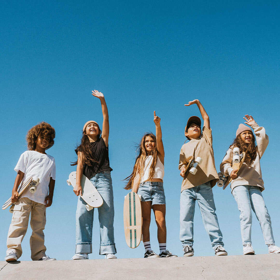 Vijf lachende kinderen staan met Banwood kinderskateboards in de hand onder een blauwe lucht.