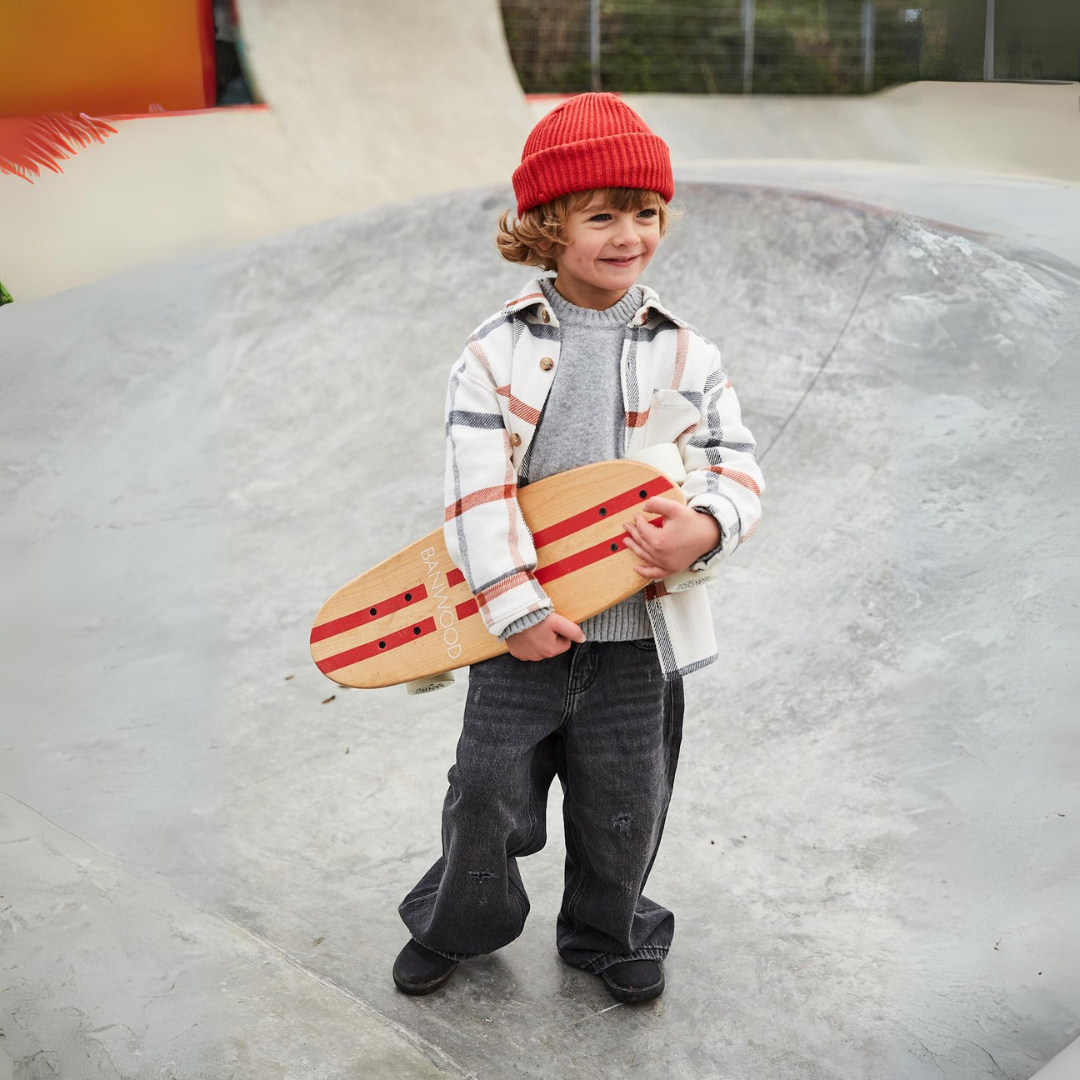 Een lachend kind met rode muts houdt het Banwood kinderskateboard vast in een skatepark.