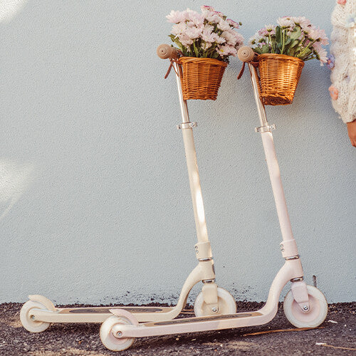 Twee Banwood kindersteps maxi in crème en roze met mandjes vol bloemen staan tegen een muur.
