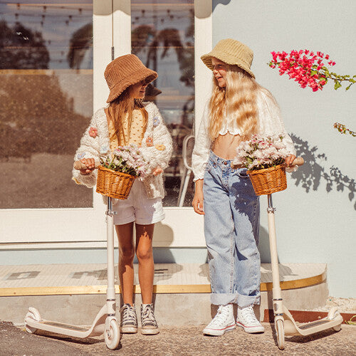 Twee meisjes met hoeden staan naast hun crème Banwood kindersteps maxi met bloemen in de mandjes.