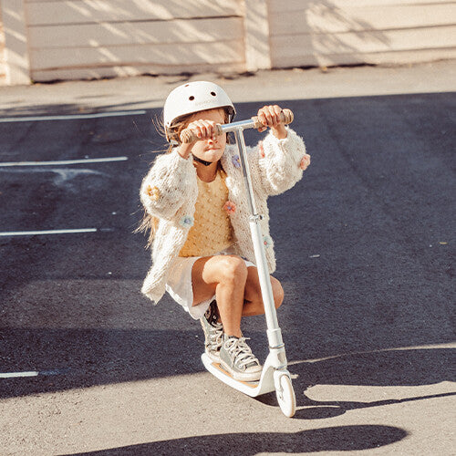 Een kind met helm hurkt op een crème Banwood kinderstep maxi met mandje op straat.
