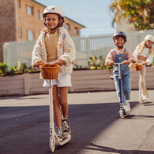Een kind met helm rijdt op een Banwood roze kinderstep met mandje, met andere kinderen op de achtergrond.