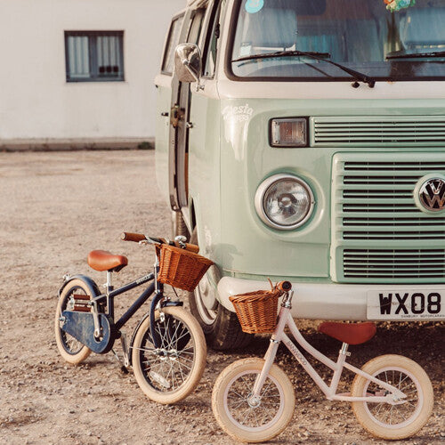 Een blauwe en een roze Banwood loopfiets met rotan mandjes staan voor een lichtgroene vintage bus.