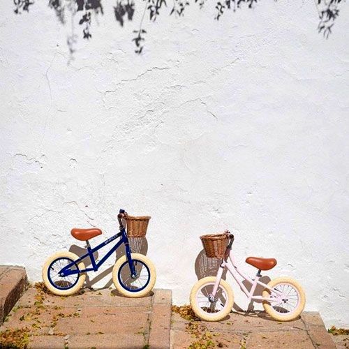 Twee Banwood loopfietsen, een blauwe en een lichtroze, staan op stenen treden tegen een witte muur.