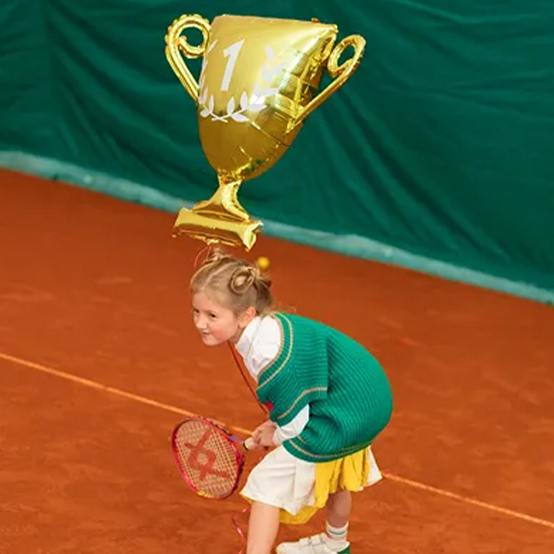 Een meisje op een tennisbaan kijkt omhoog naar de Partydeco gouden trofee folieballon die boven haar zweeft.