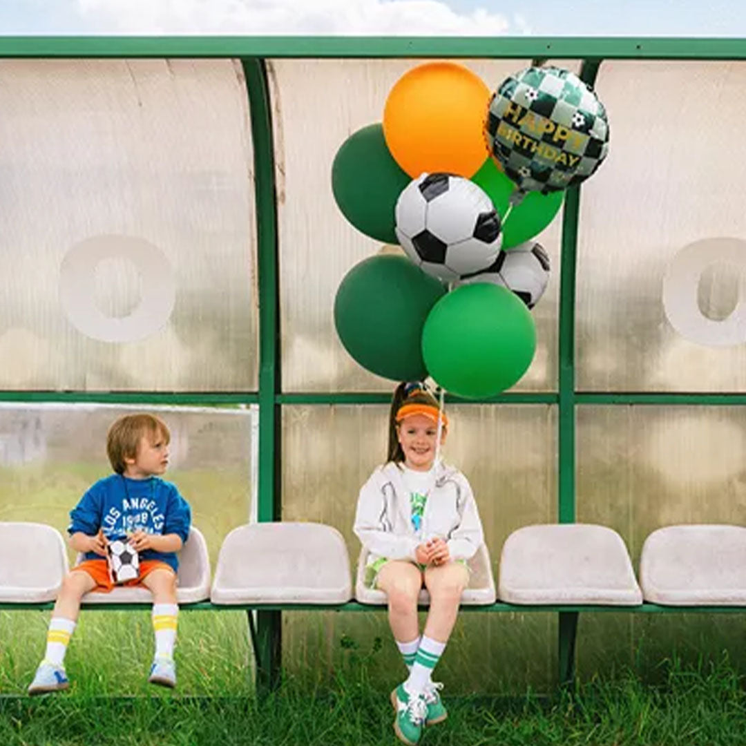 Twee kinderen zitten op een bankje in een voetbaldugout met een bos ballonnen, waaronder een groene voetbal Happy Birthday folieballon.
