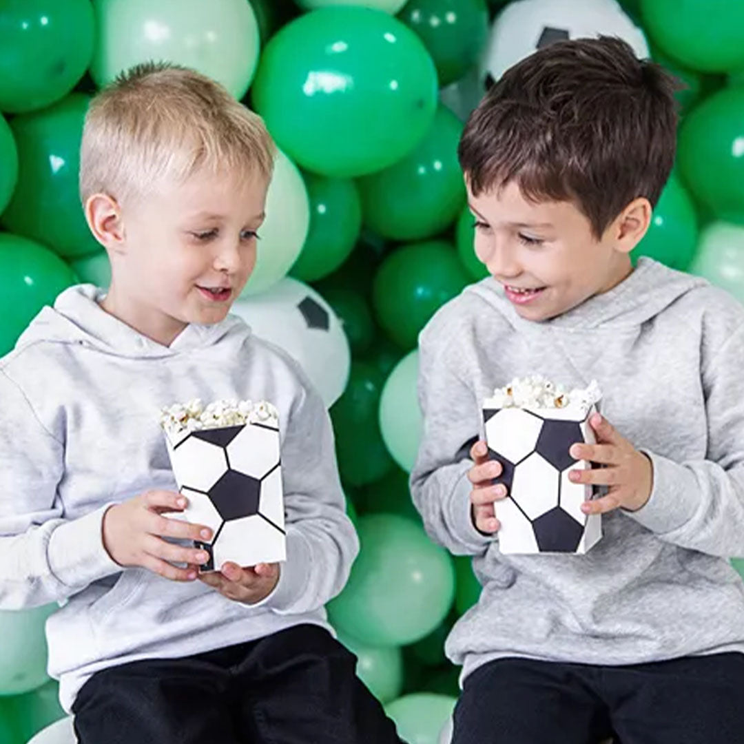 Twee jongens houden Partydeco snackbekers met voetbalpatroon en popcorn vast voor een achtergrond van groene ballonnen.