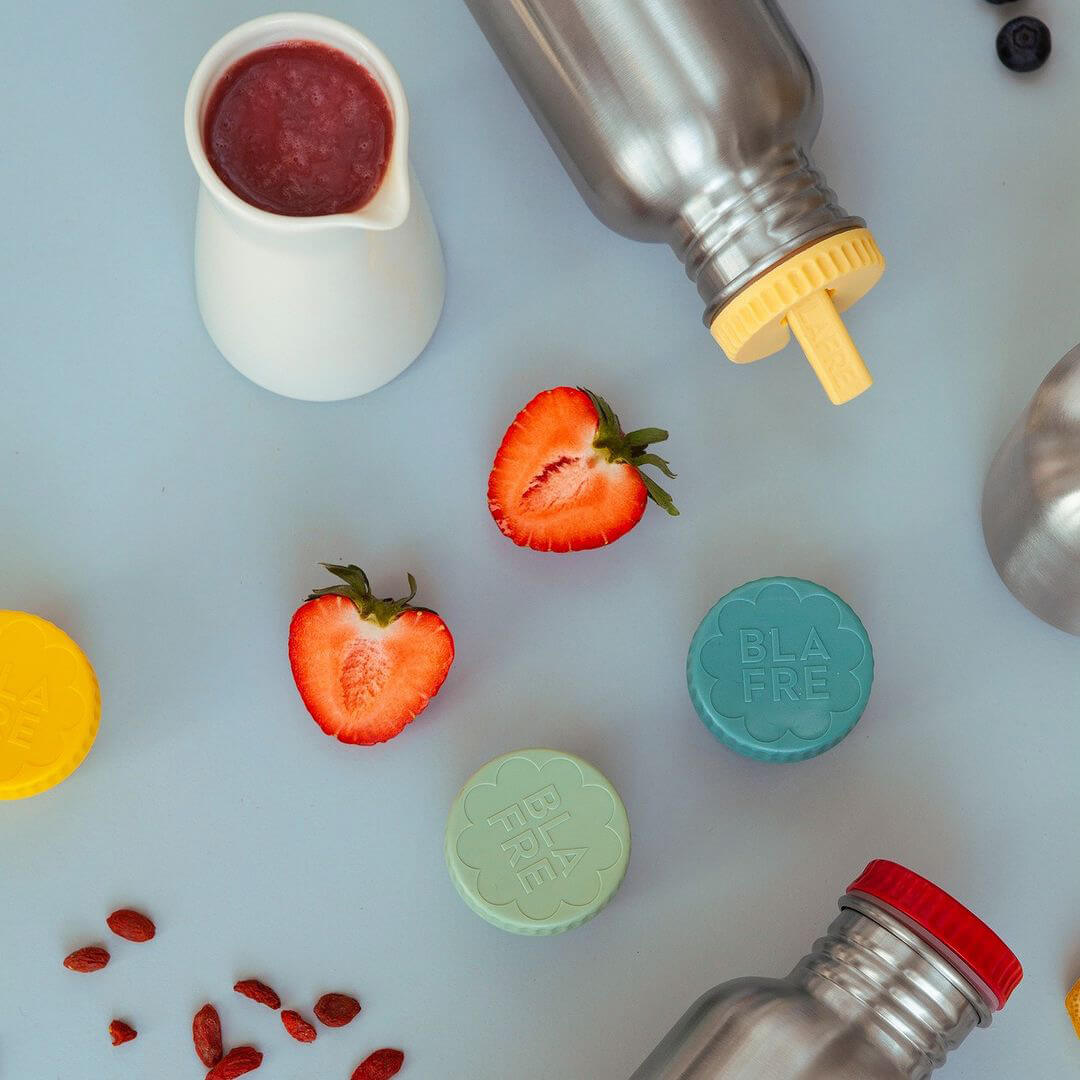 Een Blåfre drinkfles met gele drinktuit dop, omringd door aardbeien, puree en twee ronde Blåfre doppen.
