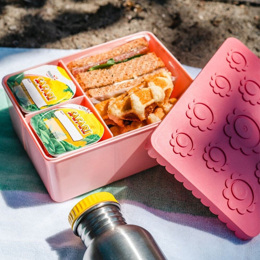 Blafre roze lunchbox gevuld met broodjes en wafels, met twee yoghurtdrankjes en een waterfles ernaast op een handdoek.