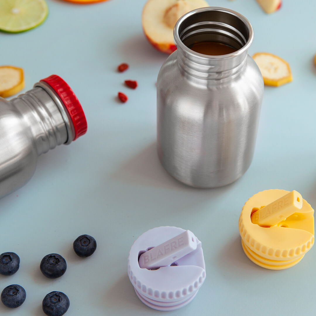 Twee Blåfre RVS drinkflessen, één met rode dop, en twee losse gekleurde drinkdoppen, omringd door bosbessen en gesneden fruit.