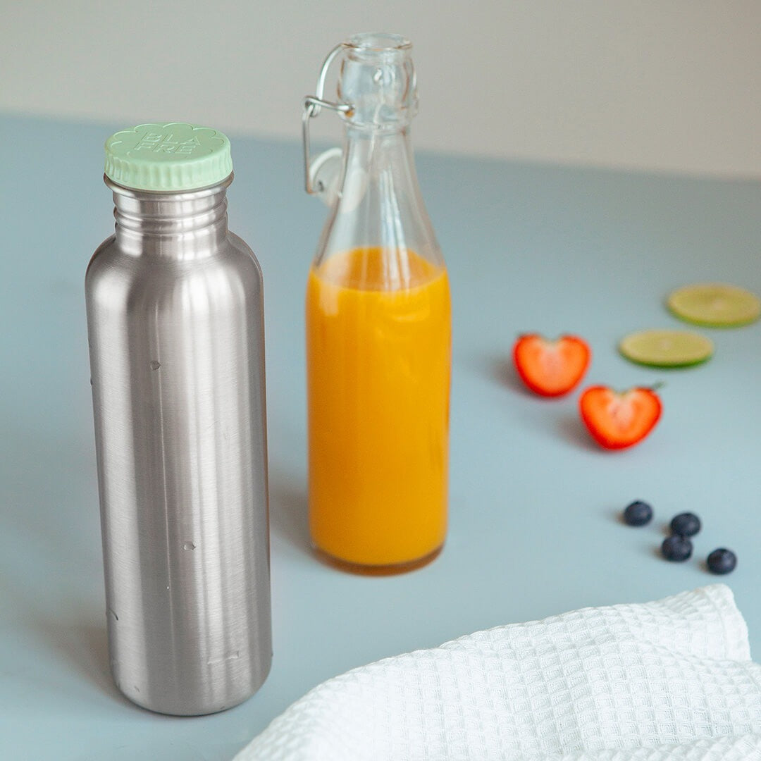 Blafre roestvrijstalen drinkfles met lichtgroene dop naast een fles met sap en vers fruit.