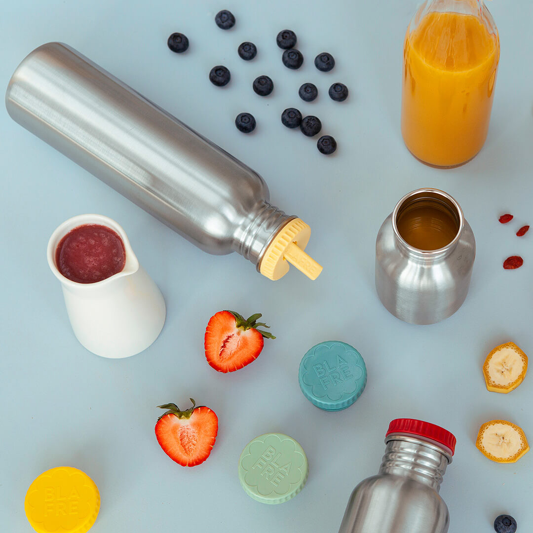 Diverse blafre RVS drinkflessen en accessoires met fruit en een kannetje puree op een lichtblauwe ondergrond.