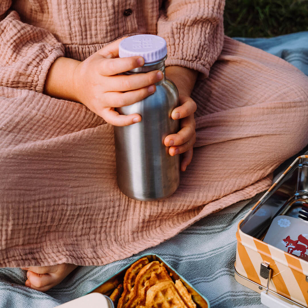 Een kind houdt de Blafre roestvrijstalen drinkfles met lila dop vast tijdens een picknick.