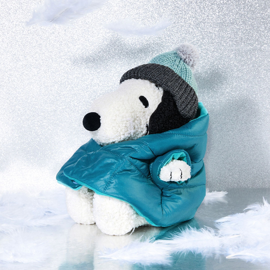 Bon Ton Toys Snoopy pluchen hond met blauwe jas en gebreide muts, zittend op een glinsterende ondergrond met witte veren.