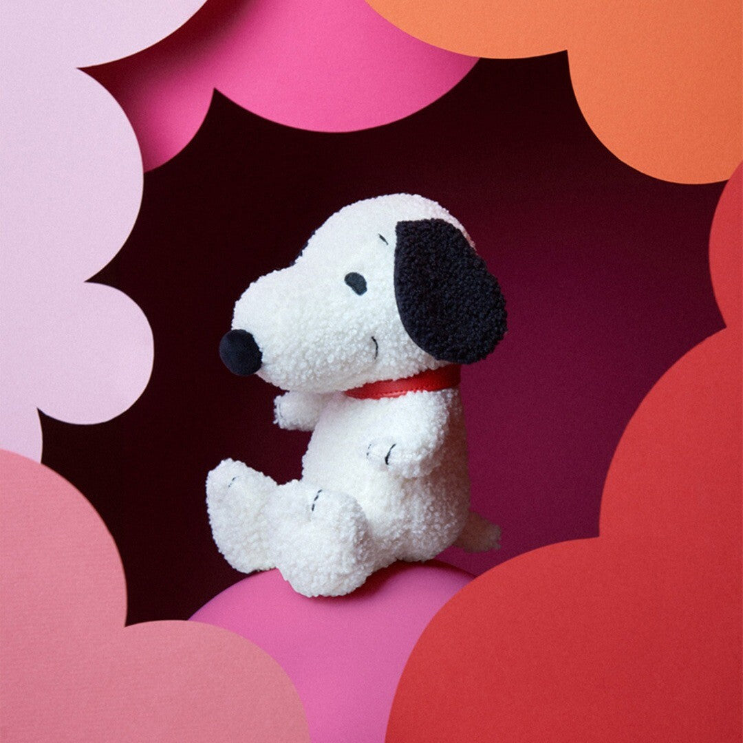Bon Ton Toys knuffelhond Snoopy zittend op een roze ondergrond, omringd door kleurrijke papieren vormen.