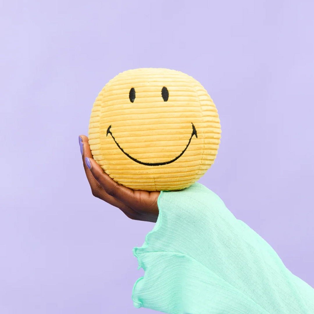 Een hand met paarse nagellak houdt de bon ton toys gele corduroy smiley knuffelbal vast tegen een paarse achtergrond.