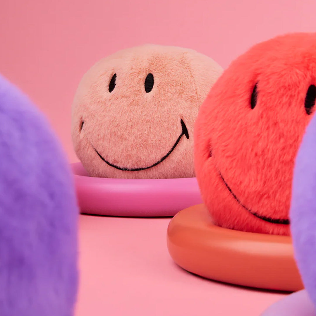Meerdere bon ton toys pluchen knuffelballen in roze, rood en paars op gekleurde standaards tegen een roze achtergrond.