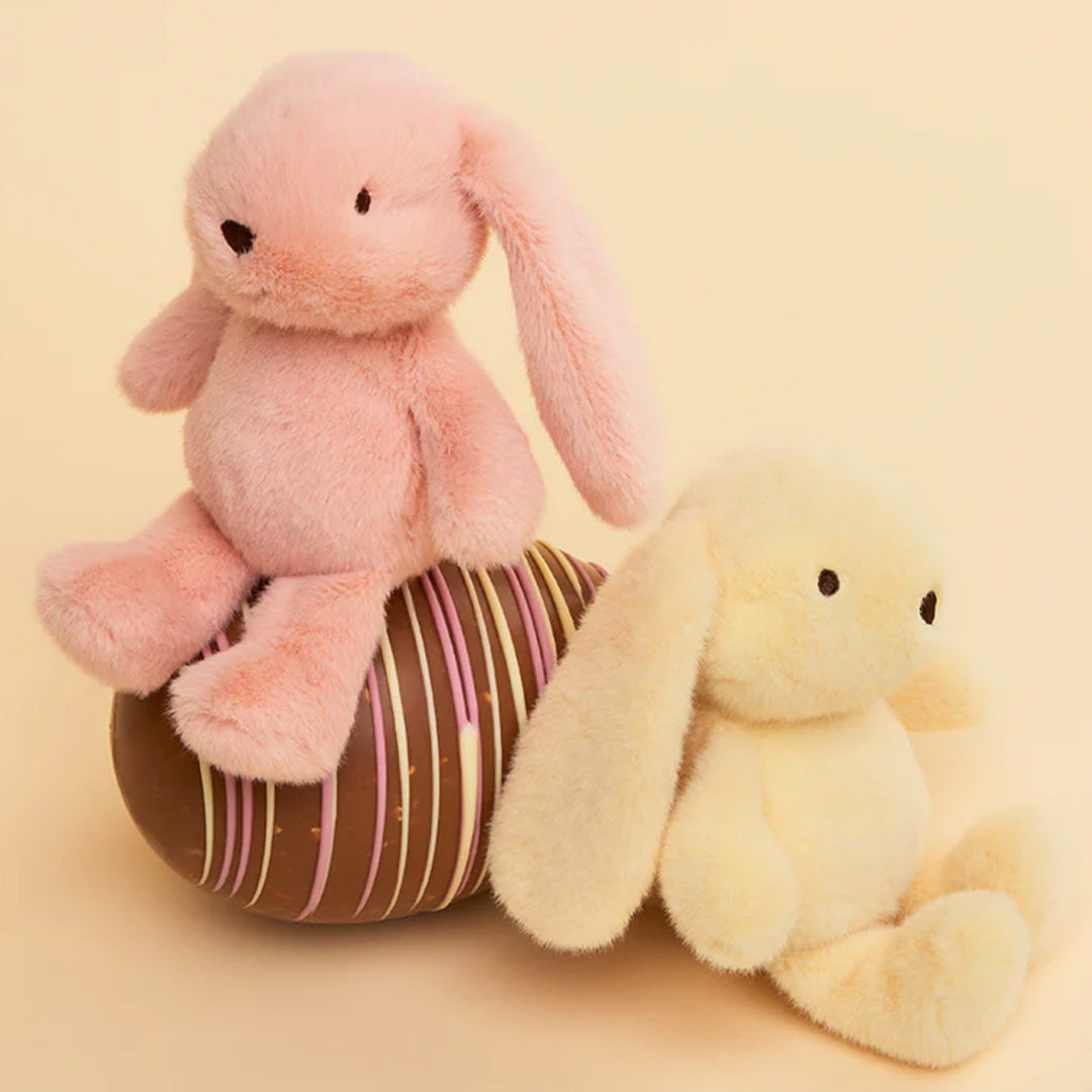 Een roze en een geel bon ton toys pluchen knuffelkonijn zitten bij een chocolade paasei.