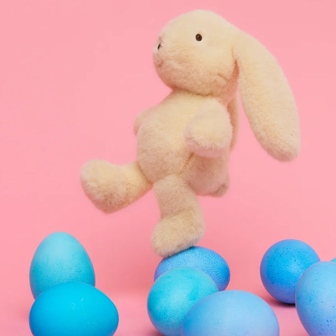 Bon ton toys geel pluchen knuffelkonijn balanceert op een blauw paasei, omringd door meer paaseieren op een roze achtergrond.