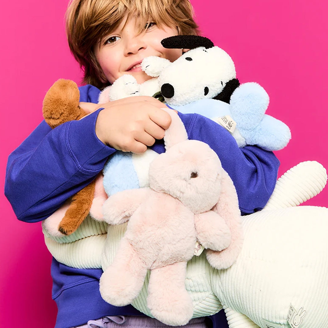 Een kind knuffelt een stapel pluchen speelgoed, waaronder het roze konijn, tegen een roze achtergrond.