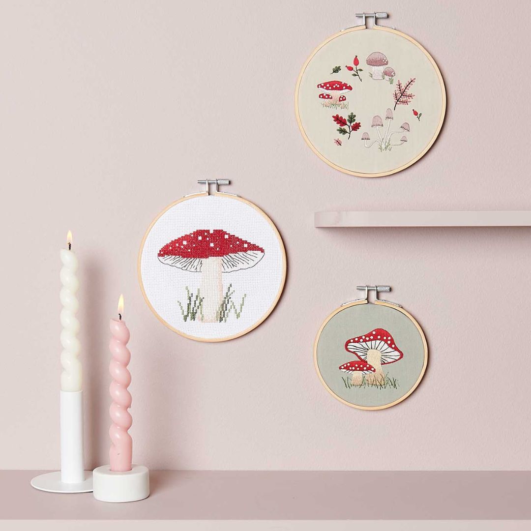 Drie geborduurde borduurringen met paddenstoelmotieven hangen aan een roze muur, naast twee kaarsen.