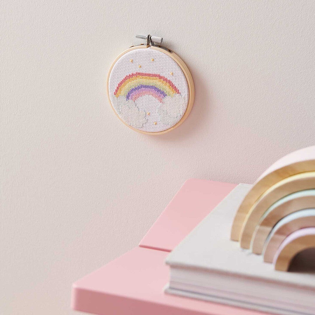 Een Rico Design kruissteek borduurwerk met regenboog en sterren hangt aan een roze muur boven een roze tafeltje.