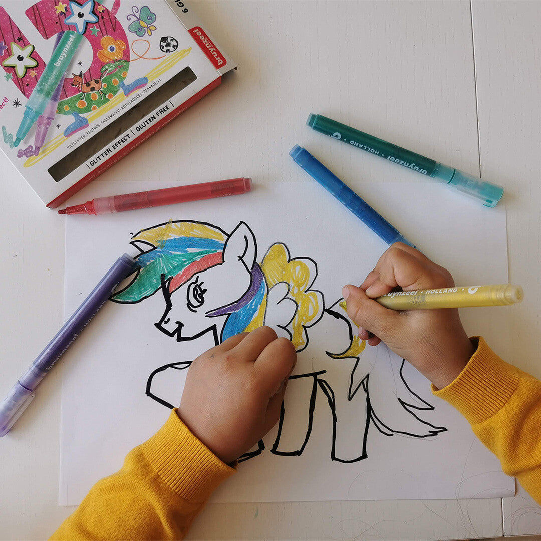 Een kind kleurt een eenhoorn met een gele Bruynzeel Kids Glitter viltstift op een witte tafel.