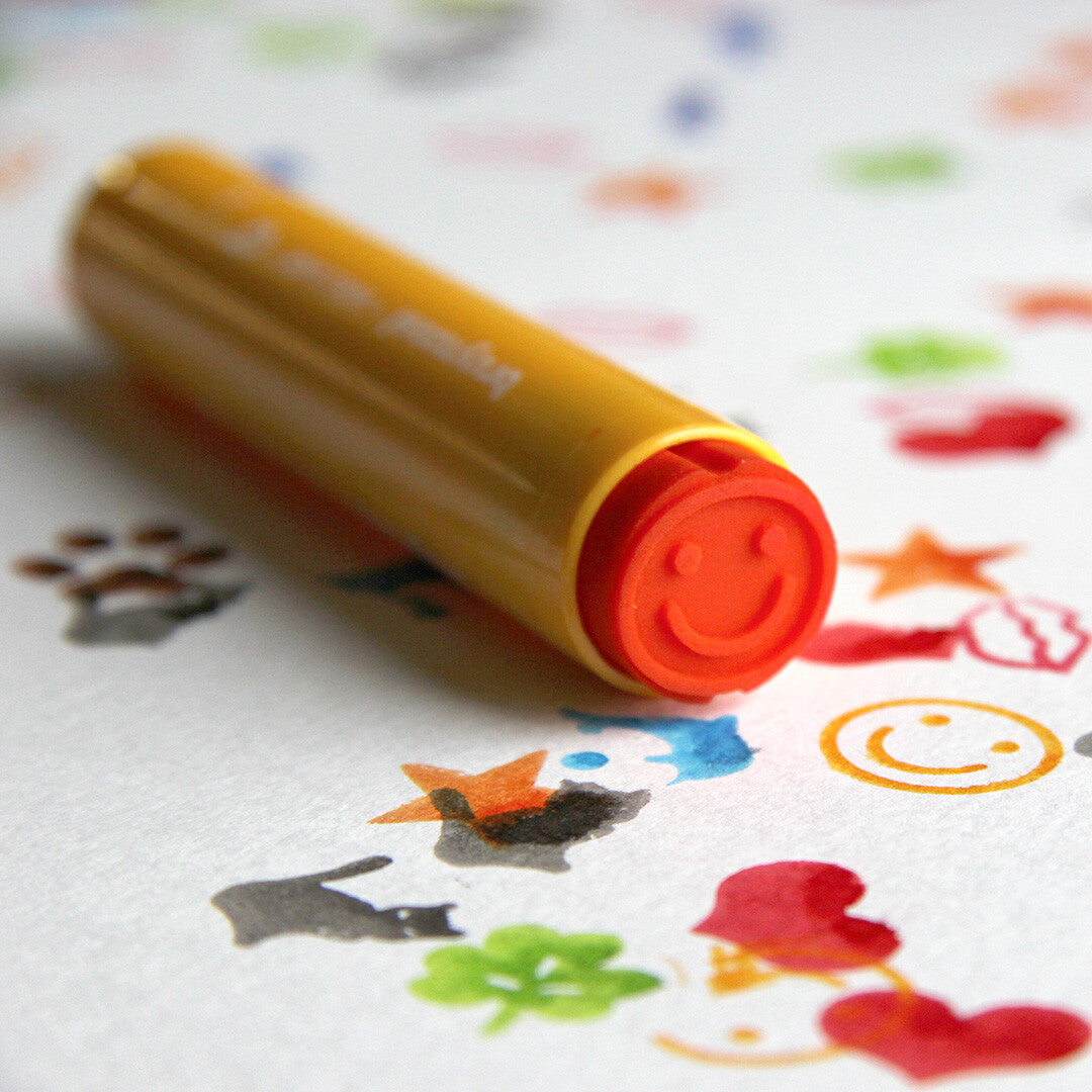 Een gele Bruynzeel stempelstift met een oranje smiley-stempel ligt tussen verschillende gestempelde figuren.
