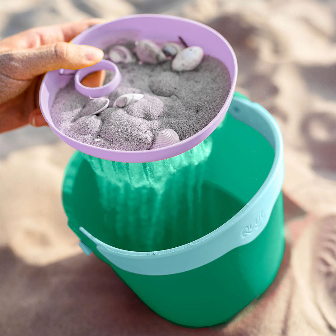 Een hand zeeft zand en schelpen met de paarse zeef boven de groene Quut strandemmer.