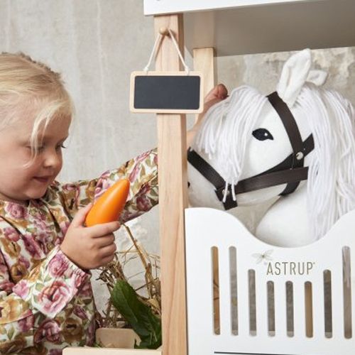 Een meisje voert een oranje speelgoedwortel aan een by astrup wit stokpaard in een houten paardenstal.