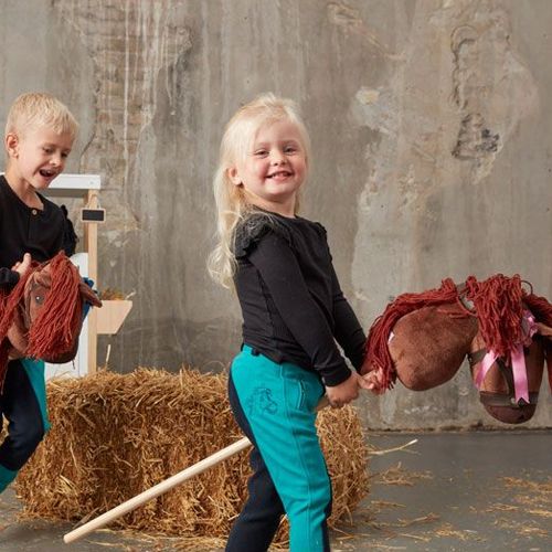 Twee lachende kinderen rennen met bruine stokpaarden in een speelomgeving.