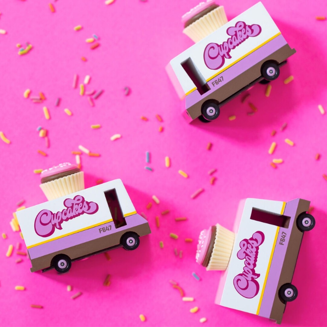 Drie Candylab speelgoed cupcake vans met cupcakes op het dak, verspreid over een roze achtergrond met gekleurde sprinkles.