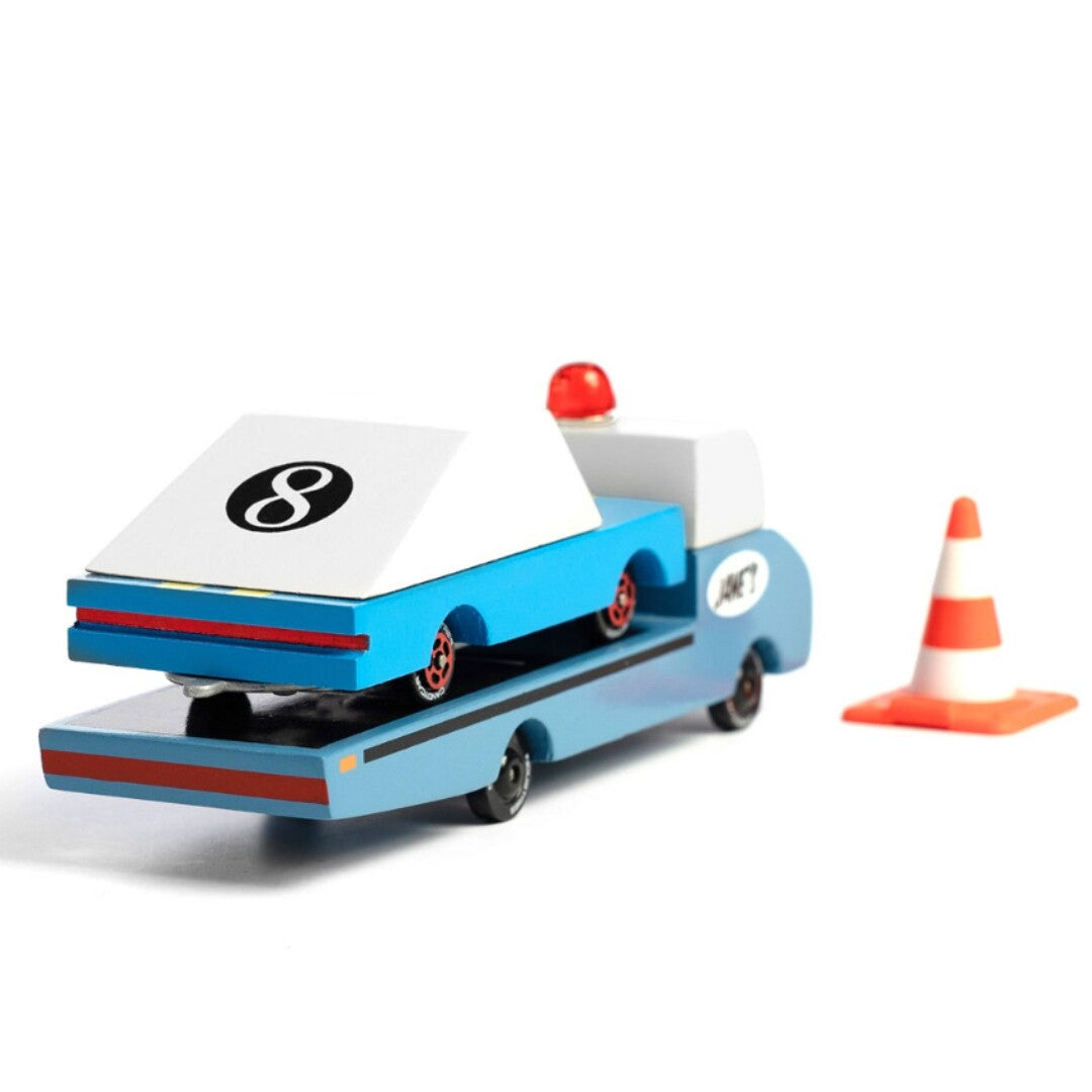 De Candylab blauwe houten speelgoedvrachtauto met een wit speelgoedautootje op de laadbak en een verkeerskegel ernaast.
