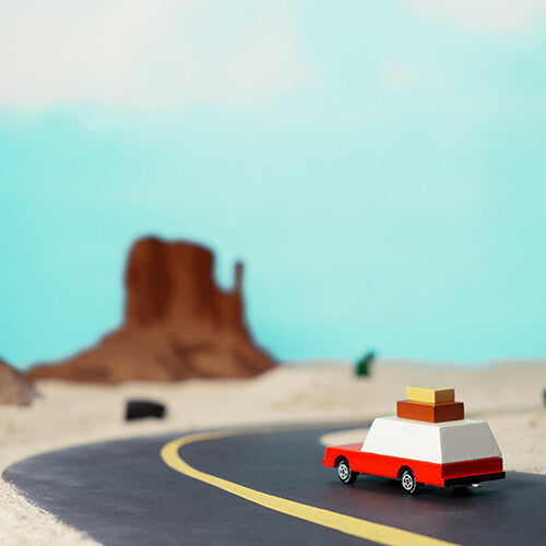 Candylab houten speelgoedauto op een weg in een woestijnlandschap onder een blauwe lucht.