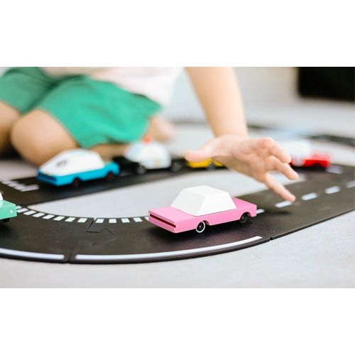 Een kinderhand reikt naar een Candylab roze houten speelgoedauto op een zwarte speelgoedweg.